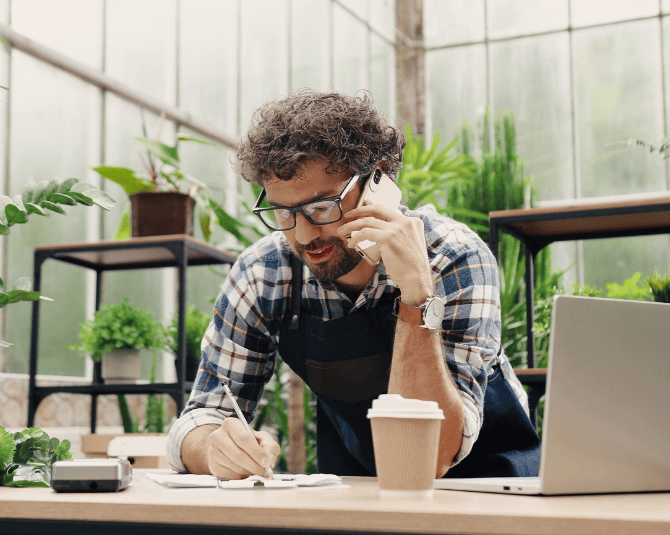 Man on phone at the counter with his laptop in front of shelves full of plants.
